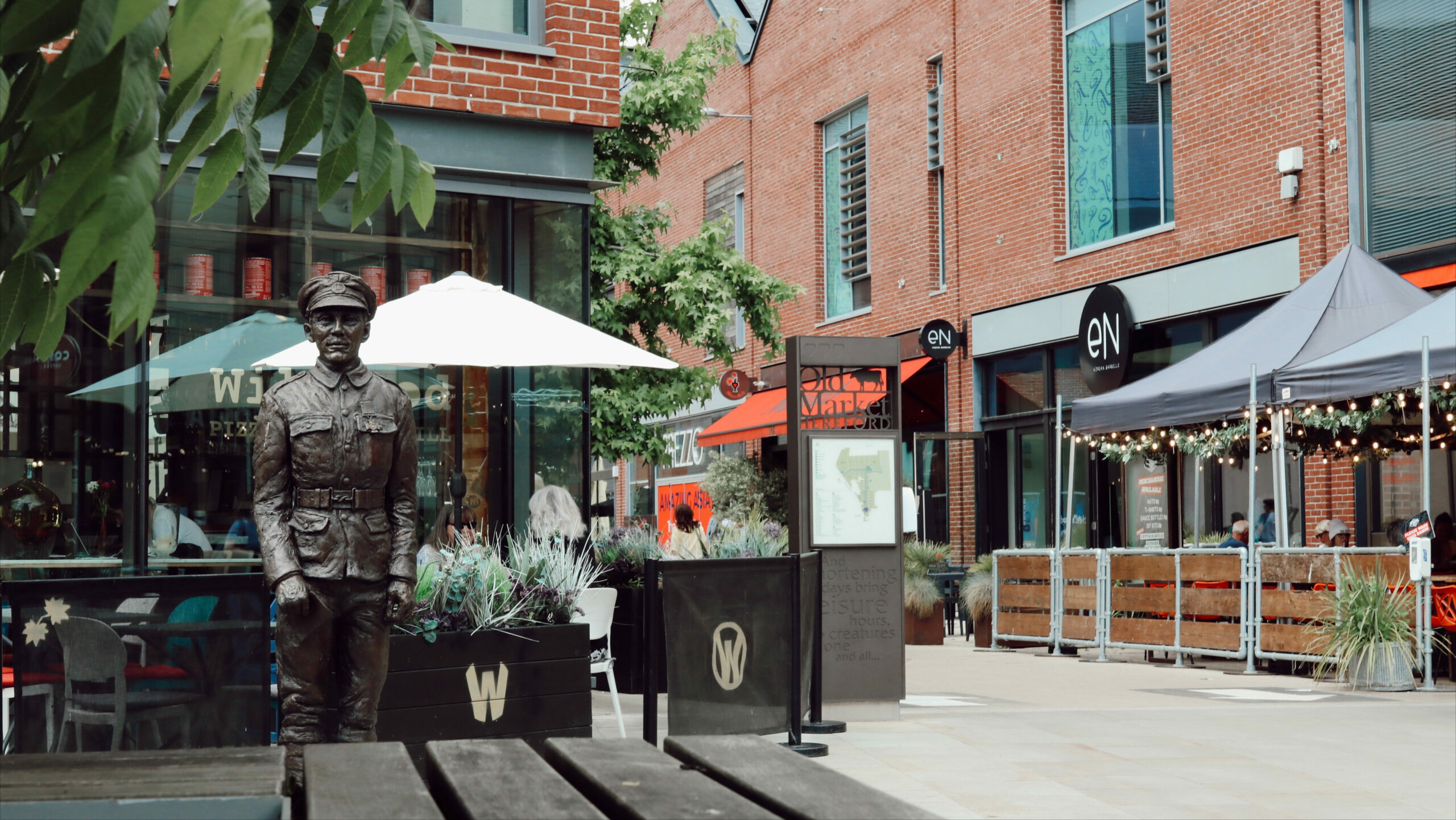 Statue of a soldier in a modern shopping and dining area in Hereford, with outdoor seating, shops, and cafes in the background.