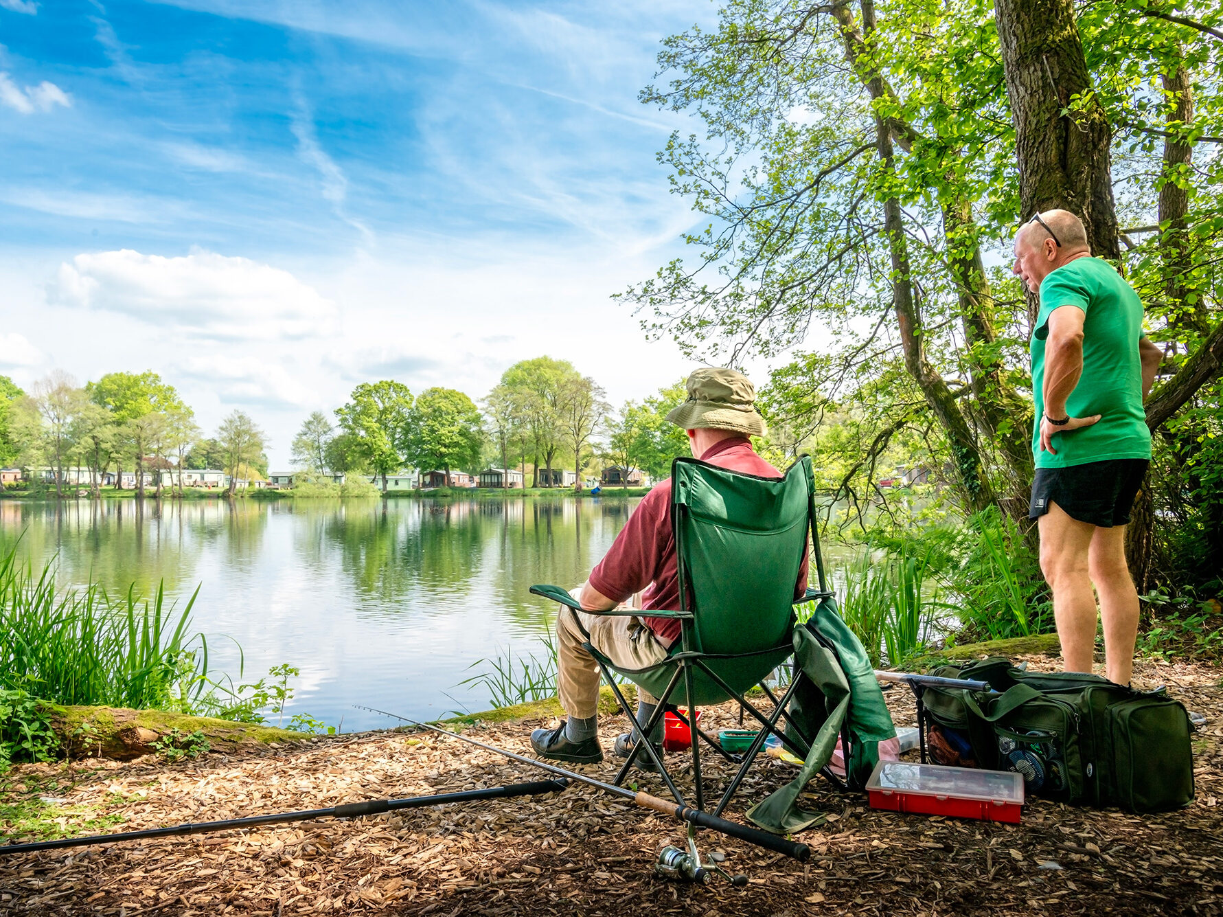 Two men fishing at Pearl Lake in Herefordshire, sitting beside calm water surrounded by trees and blue sky at Discover Parks.